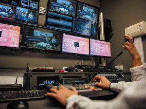 2 peoples hands only on keyboard / control panel in TV production room, with multiple screens in front of them.