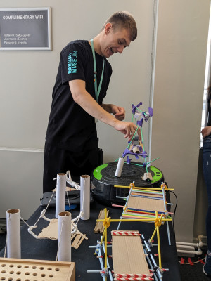 Male member of National Railway Museum staff smiling and holding a student built bridge (made out of tape, straws and cardboard)