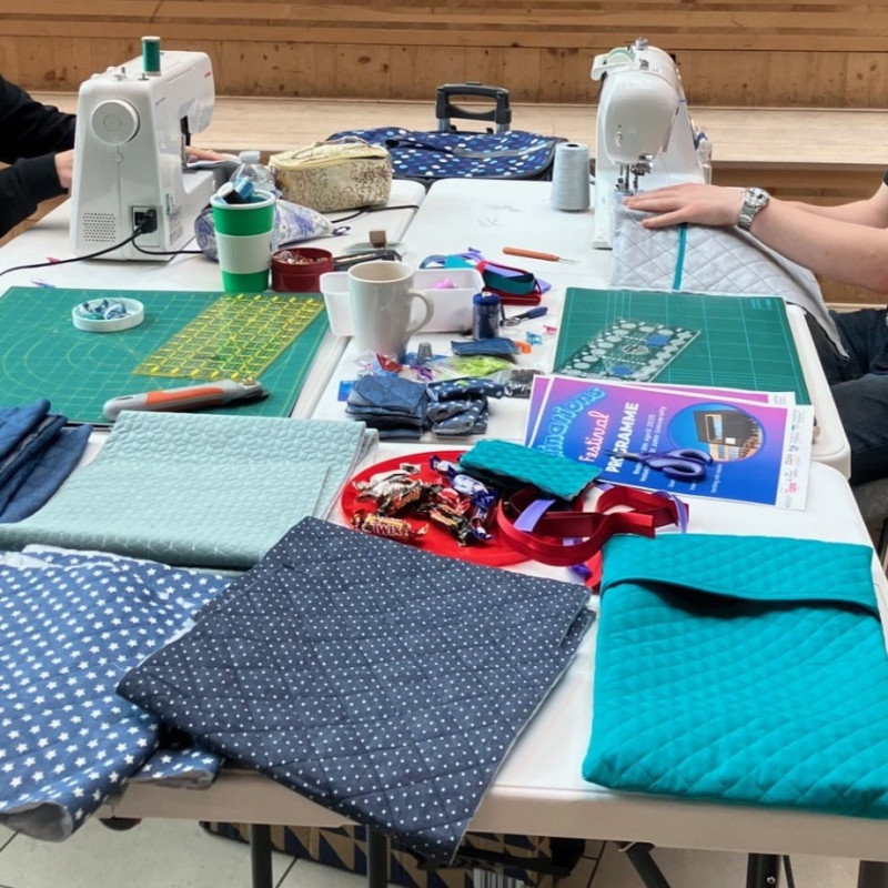 Two pairs of hands sewing at sewing machines on a table filled with sewing materials.
