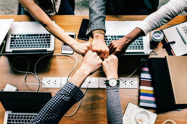 Five people fist-bumping over their work station and laptops